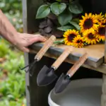 3-piece iron garden tool set with wooden handles: small rake, wide trowel, narrow trowel resting on a wooden shelf near sunflowers