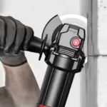 Gloved hand operating a black brushless angle grinder with a red button, cutting a gray metal workpiece