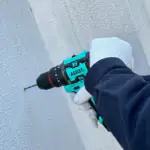 First-person view of a gloved hand using a teal ASSIST cordless impact drill to drill into a textured white wall