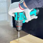 Gloved worker drives a screw into light wood using a teal ASSIST cordless impact drill, with an indoor workshop background