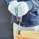 Gloved worker uses a teal 12V lithium battery impact drill to bore a hole in a light wood block, producing wood shavings