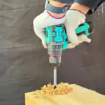 Gloved hands holding a teal 12V lithium battery impact drill drilling into wood, with curled wood shavings around the bit