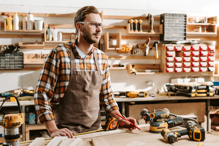 Woodworker in safety glasses and apron marks lumber at a workbench surrounded by yellow and black power tools.