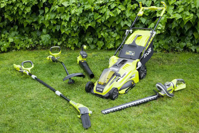 Bright yellow and black RYOBI cordless garden tool set laid out on green grass in front of a leafy hedge.