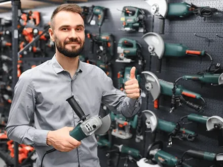 Bearded man in a gray shirt holds a corded angle grinder and gives a thumbs up in front of a pegboard wall of power tools