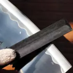 Close-up of a gloved hand holding a black abrasive diamond file sharpening a shiny metal blade edge.